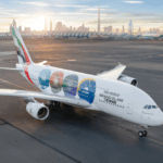 An Emirates Airbus A380-800 parked on the tarmac at Dubai International Airport at sunrise. The aircraft features a special "Grand Slam Tennis" livery with four colorful tennis ball icons representing the Australian Open, Roland-Garros, Wimbledon, and the US Open. The Dubai skyline, featuring the Burj Khalifa, is visible in the background under a soft morning sky.