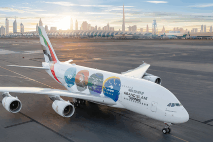 An Emirates Airbus A380-800 parked on the tarmac at Dubai International Airport at sunrise. The aircraft features a special "Grand Slam Tennis" livery with four colorful tennis ball icons representing the Australian Open, Roland-Garros, Wimbledon, and the US Open. The Dubai skyline, featuring the Burj Khalifa, is visible in the background under a soft morning sky.