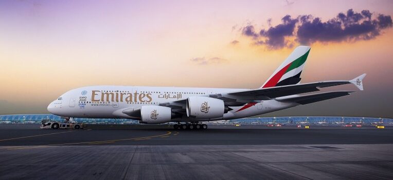 An Emirates Airbus A380 is parked on an airport apron during a vibrant purple and orange sunset. The aircraft is shown in a side profile, prominently displaying the 