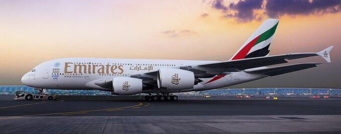 An Emirates Airbus A380 is parked on an airport apron during a vibrant purple and orange sunset. The aircraft is shown in a side profile, prominently displaying the 