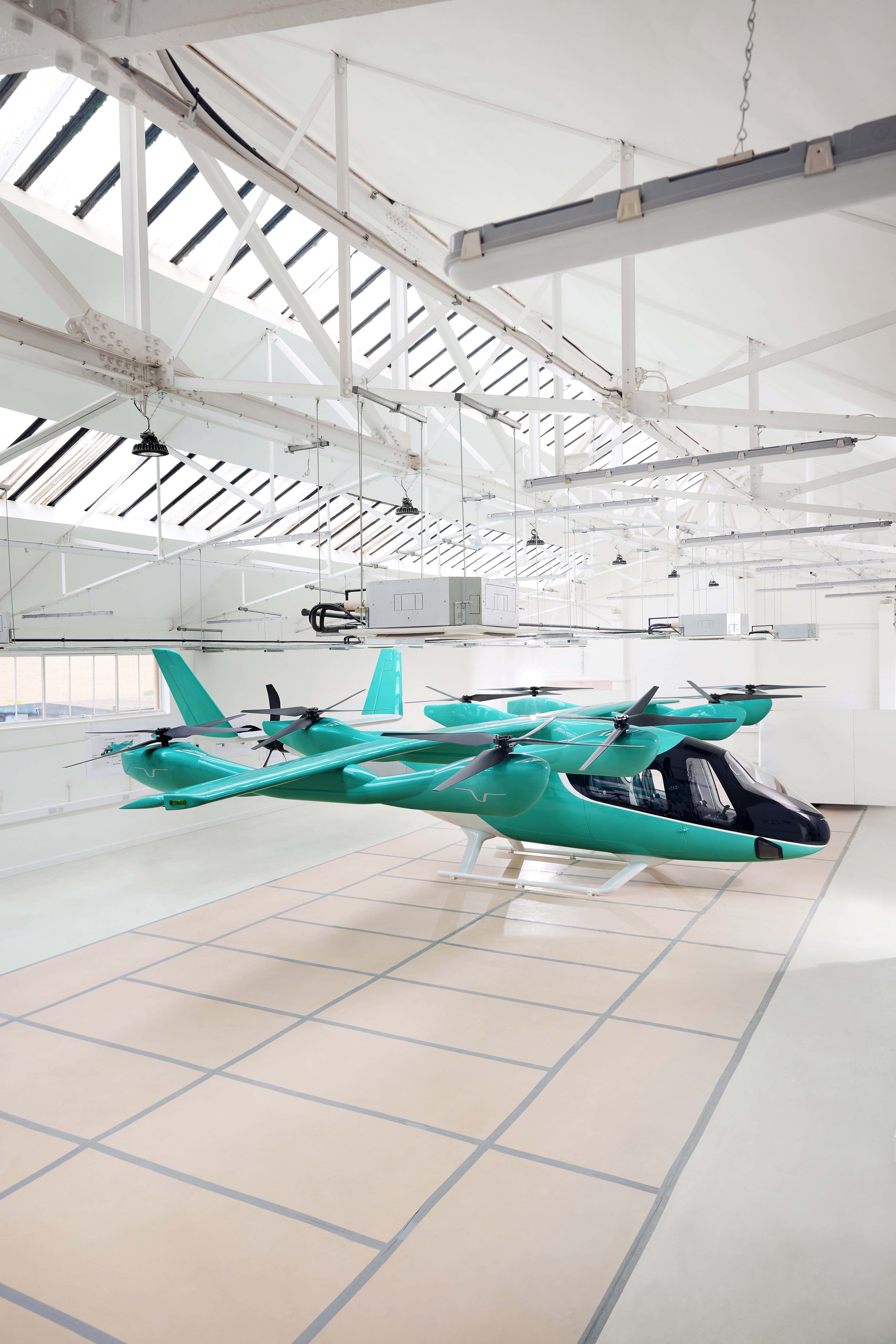 A side-profile view of a teal and white electric vertical takeoff and landing (eVTOL) aircraft parked inside a bright, white industrial hangar. The aircraft features a sleek, aerodynamic fuselage, a large black cockpit canopy, and eight horizontal rotors mounted on two long parallel wings for vertical lift. A traditional tail fin is visible at the rear, and the craft rests on white landing skids over a grid-patterned floor.