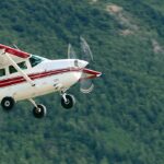 A white and red Cessna U206F Stationair, registration N74PS, captured in mid-flight against a blurred background of a dense, green evergreen forest. The single-engine aircraft is banked slightly, showing its fixed landing gear and rotating propeller. The logo for "Kantishna Air Taxi" is visible on the tail.