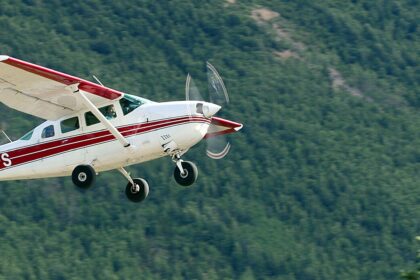 A white and red Cessna U206F Stationair, registration N74PS, captured in mid-flight against a blurred background of a dense, green evergreen forest. The single-engine aircraft is banked slightly, showing its fixed landing gear and rotating propeller. The logo for "Kantishna Air Taxi" is visible on the tail.