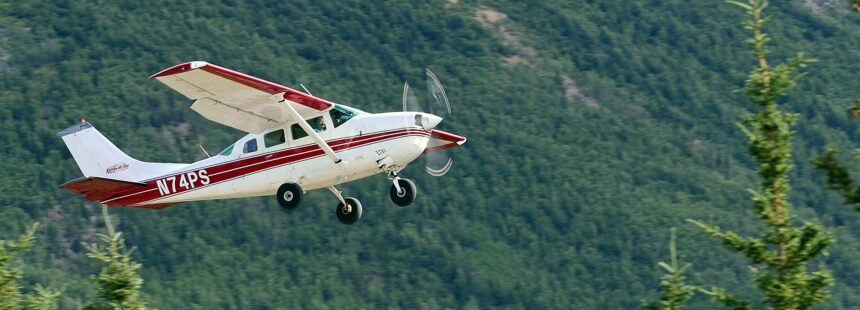 A white and red Cessna U206F Stationair, registration N74PS, captured in mid-flight against a blurred background of a dense, green evergreen forest. The single-engine aircraft is banked slightly, showing its fixed landing gear and rotating propeller. The logo for "Kantishna Air Taxi" is visible on the tail.