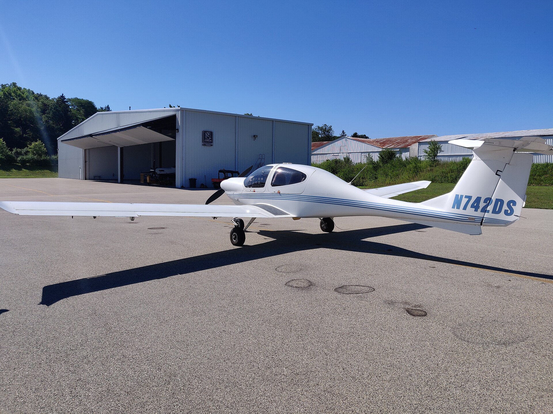 A sleek, white Diamond DA20-C1 Katana light aircraft, registration N742DS, is parked on an asphalt apron under a clear blue sky. The two-seat aircraft features a low-wing design, a bubble canopy, and a T-tail. In the background, a large white hangar with its door open and several smaller buildings are visible.