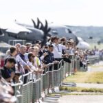 A large crowd of people stands along a metal security fence at an airfield. Many individuals are holding up smartphones to take pictures of large military transport aircraft, including one with prominent dark propellers, visible in the blurred background. The setting is outdoors on a clear day with a paved taxiway and grassy verge in the foreground.