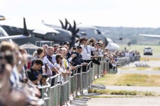 A large crowd of people stands along a metal security fence at an airfield. Many individuals are holding up smartphones to take pictures of large military transport aircraft, including one with prominent dark propellers, visible in the blurred background. The setting is outdoors on a clear day with a paved taxiway and grassy verge in the foreground.