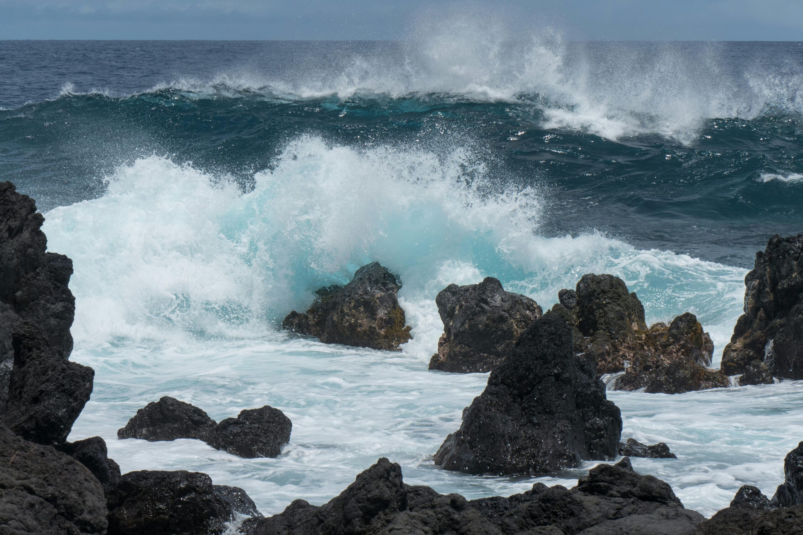 Powerful turquoise waves crashing against a rugged shoreline of jagged, black volcanic rocks. The white ocean spray and foam contrast sharply against the dark stones and the deep blue of the Pacific Ocean under an overcast sky.