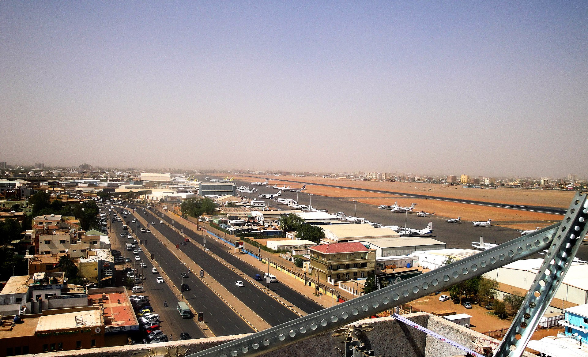 A high-angle, wide-reaching view of Khartoum International Airport in Sudan. To the left, a busy multi-lane highway is filled with cars next to urban buildings. To the right, the airport runway and tarmac are visible, lined with various aircraft and hangars under a hazy, pale sky.