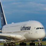 A Lufthansa aircraft sits on the runway under a blue sky, its bright white fuselage clearly visible