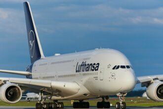 A Lufthansa aircraft sits on the runway under a blue sky, its bright white fuselage clearly visible