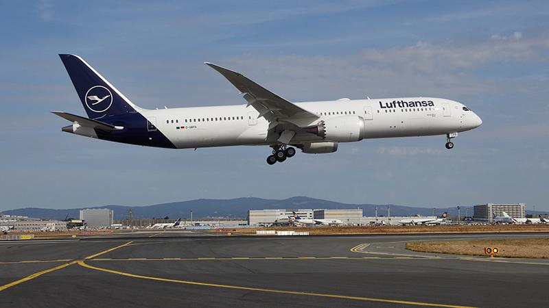 A Lufthansa aircraft descends and lands on the tarmac under a clear sky, with runway details visible below.
