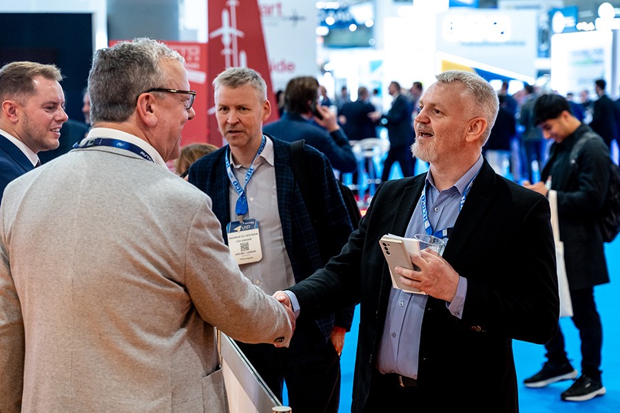 Two men in business casual attire shake hands on a bright blue carpeted floor at the MRO Europe exhibition in Barcelona, Spain. One man wears a tan blazer, while the other wears a dark blazer and a blue lanyard. In the background, other attendees and various exhibition booths are visible, including a red vertical banner with an aircraft graphic. The setting is a large, brightly lit convention hall.
