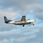 A white and navy blue Mokulele Airlines Cessna 208B Grand Caravan EX aircraft flying through a light blue sky with soft, scattered clouds. The plane features the "MOKULELE" logo on its fuselage and a stylized floral design on its tail. The landing gear is deployed as it appears to be in its landing or takeoff phase.