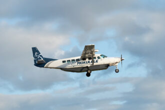 A white and navy blue Mokulele Airlines Cessna 208B Grand Caravan EX aircraft flying through a light blue sky with soft, scattered clouds. The plane features the "MOKULELE" logo on its fuselage and a stylized floral design on its tail. The landing gear is deployed as it appears to be in its landing or takeoff phase.