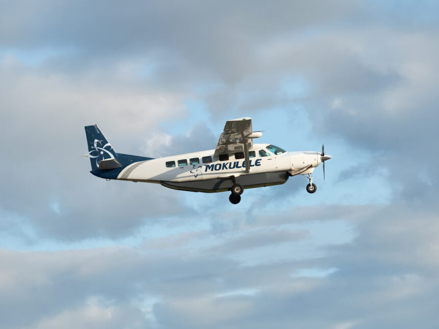 A white and navy blue Mokulele Airlines Cessna 208B Grand Caravan EX aircraft flying through a light blue sky with soft, scattered clouds. The plane features the "MOKULELE" logo on its fuselage and a stylized floral design on its tail. The landing gear is deployed as it appears to be in its landing or takeoff phase.