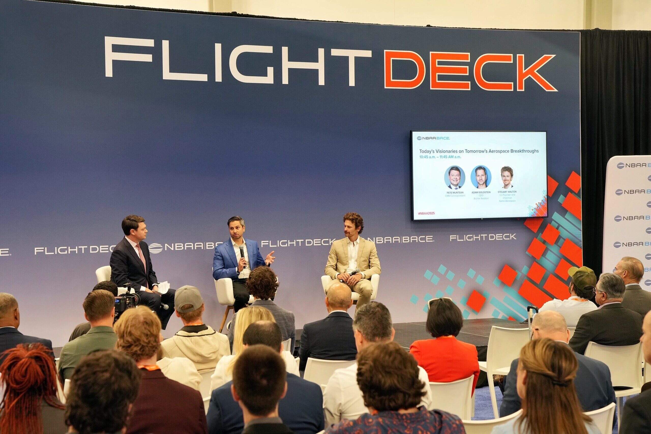 A wide shot of a stage set up for a presentation. Three men sit in white chairs during a panel discussion in front of a large blue backdrop that says 