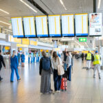 Amsterdam Airport Schiphol departure hall. Passengers carry suitcases and look at the gate screens.