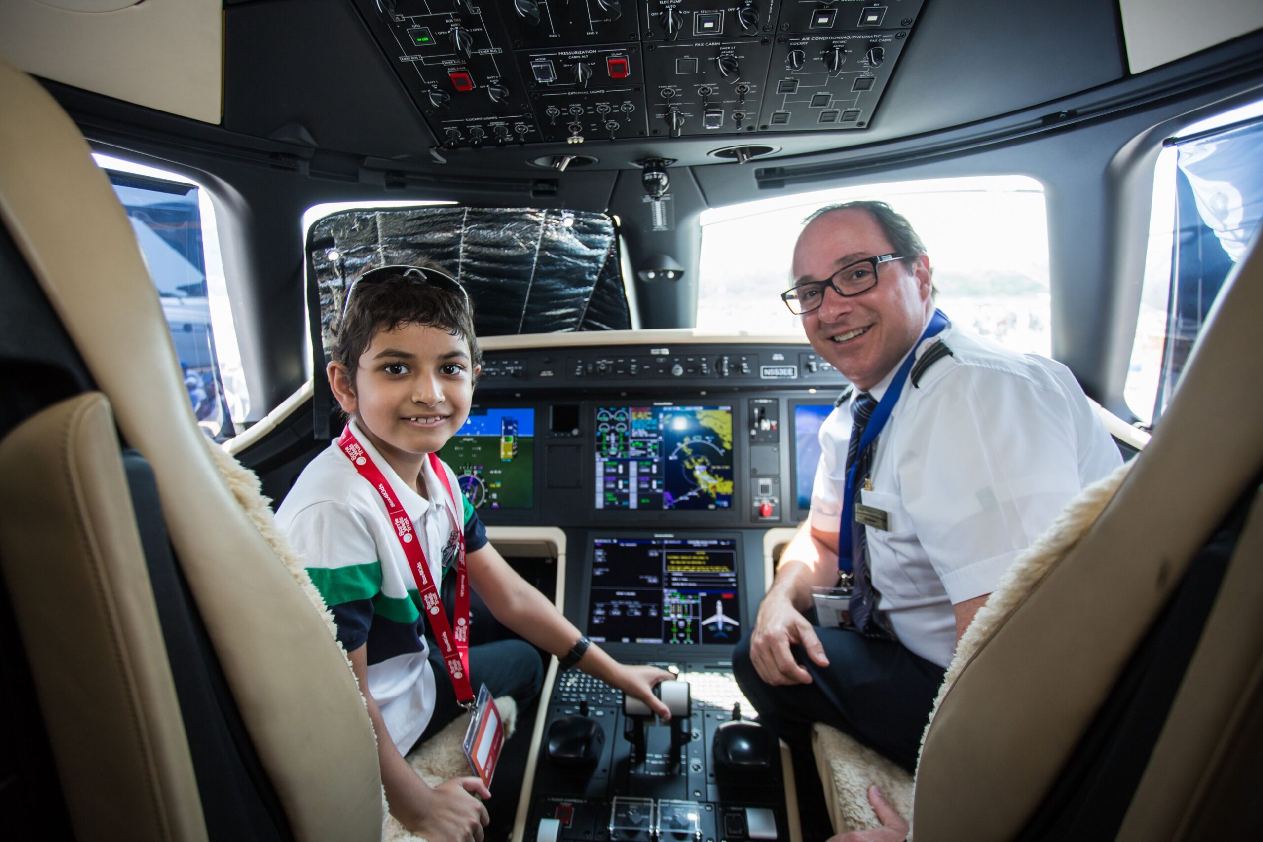 A young boy and a smiling pilot sit in the cockpit of a modern business jet. The boy, wearing a white and green shirt with a red lanyard, sits in the left pilot's seat with his hand on the throttle. The pilot sits in the right seat, wearing a white uniform and glasses. The background features complex digital instrument displays (glass cockpit), overhead panels, and plush tan leather seating.