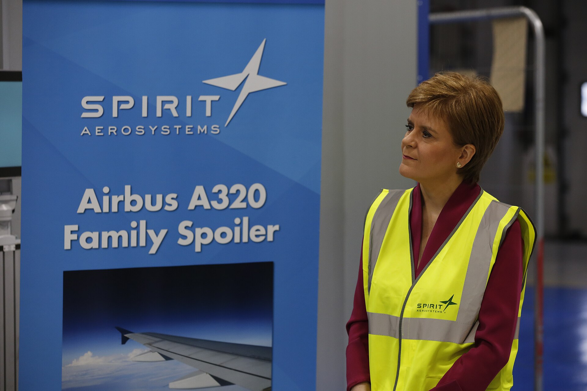 A medium shot of Nicola Sturgeon, former First Minister of Scotland, standing in an industrial setting. She is wearing a maroon blouse and a bright yellow high-visibility safety vest with the Spirit AeroSystems logo. To her left is a large blue banner that reads 