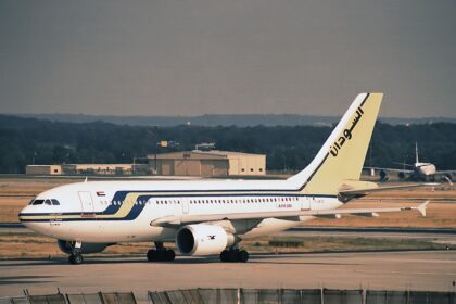 A Sudan Airways Airbus A310-300 sits on an airport tarmac. The plane features a white fuselage with blue and yellow stripes and Arabic calligraphy on the tail.