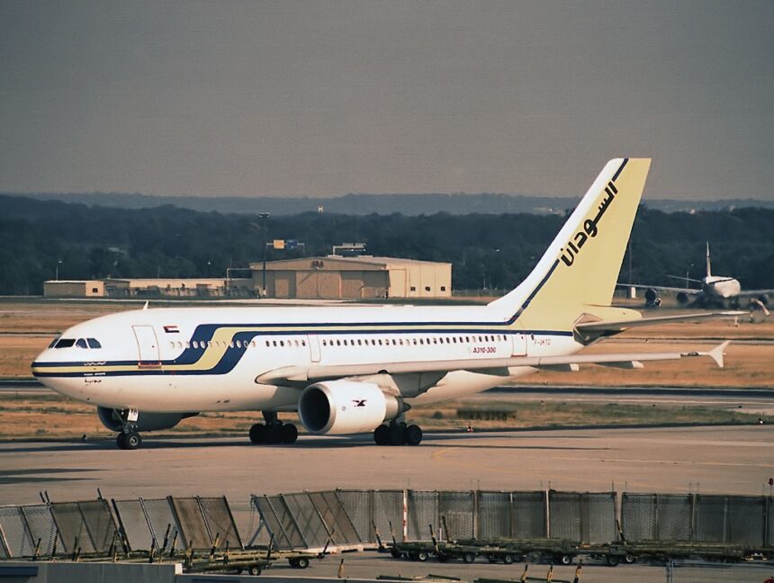 A Sudan Airways Airbus A310-300 sits on an airport tarmac. The plane features a white fuselage with blue and yellow stripes and Arabic calligraphy on the tail.