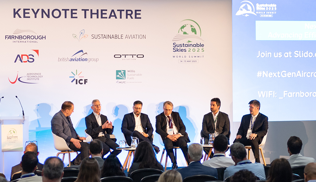 A panel of six speakers sits on a stage in the "Keynote Theatre" during the Sustainable Skies World Summit 2025. The panel consists of six men in business attire sitting in white chairs, with one speaker actively gesturing while talking into a microphone. Behind them is a large white backdrop featuring logos for Farnborough International, Sustainable Aviation, ADS, British Aviation Group, Otto Aviation, Aerospace Technology Institute, ICF, and Willis Sustainable Fuels. A large screen to the right displays event information, including a Slido link and the hashtag #NextGenAircraft. An audience is visible in the foreground, viewed from behind.