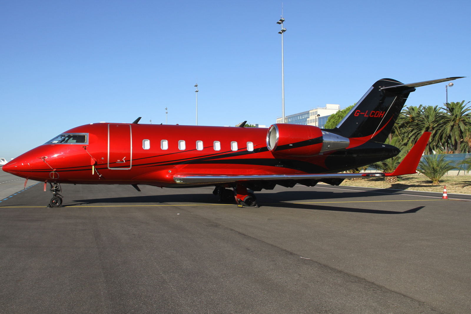 A vibrant metallic red private jet with black sweeping accents parked on an asphalt tarmac. The aircraft, a Bombardier Challenger 605 with registration G-LCDH, features a T-tail and large winglets. Palm trees and a building are visible in the background under a clear sky.
