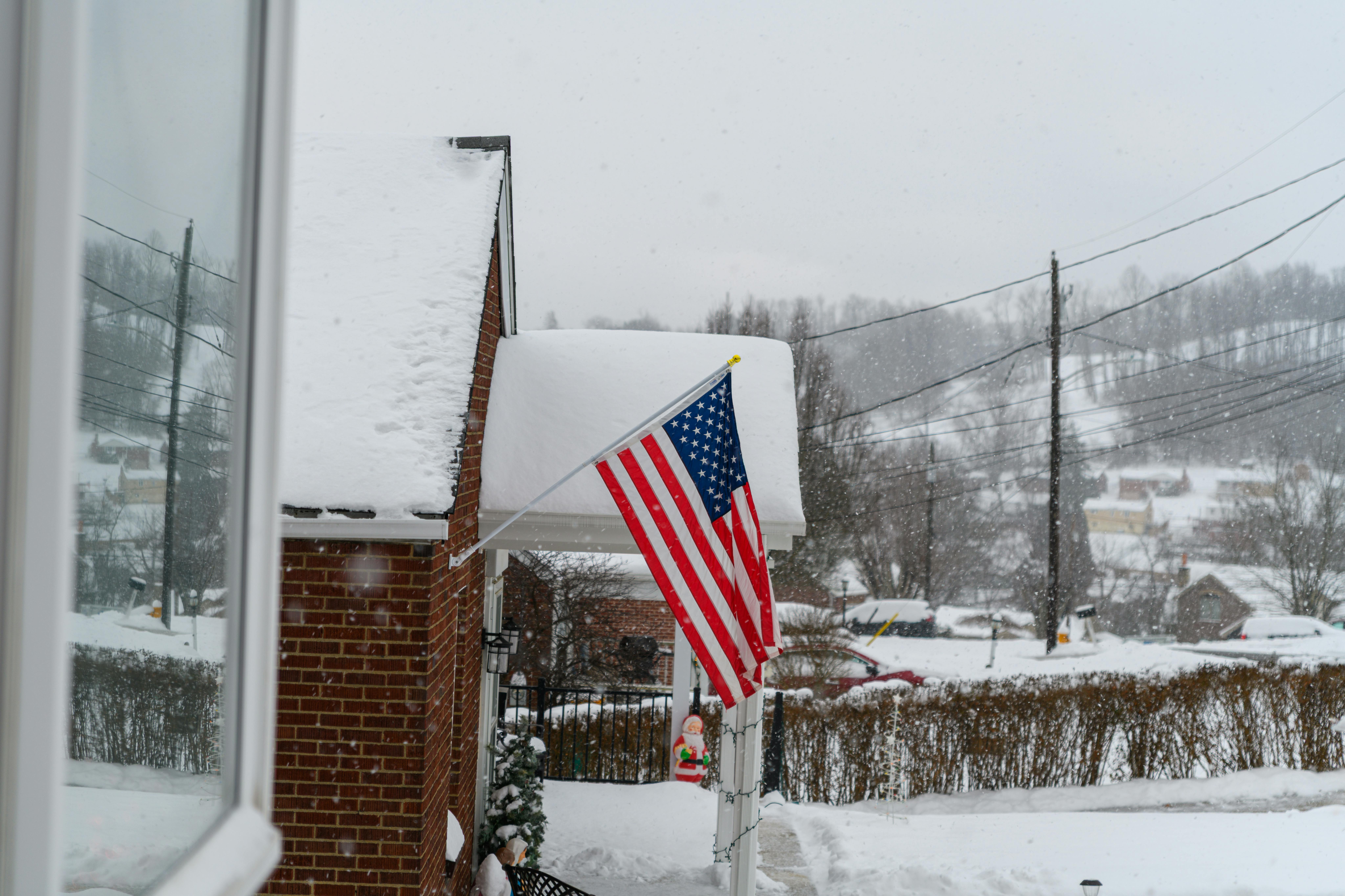 An American flag hangs from a snow-covered brick house during a winter storm. The surrounding neighborhood and hilly background are blanketed in thick white snow under a grey, overcast sky.