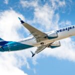 A low-angle view of a WestJet Boeing 737 MAX 8 climbing into a bright blue sky filled with white, fluffy clouds. The aircraft displays the modern WestJet livery, featuring a white upper fuselage with teal and dark blue accents toward the rear and tail. The registration C-FNWD is visible on the fuselage, and the plane's gear is retracted as it ascends.