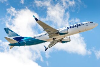 A low-angle view of a WestJet Boeing 737 MAX 8 climbing into a bright blue sky filled with white, fluffy clouds. The aircraft displays the modern WestJet livery, featuring a white upper fuselage with teal and dark blue accents toward the rear and tail. The registration C-FNWD is visible on the fuselage, and the plane's gear is retracted as it ascends.