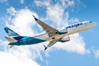 A low-angle view of a WestJet Boeing 737 MAX 8 climbing into a bright blue sky filled with white, fluffy clouds. The aircraft displays the modern WestJet livery, featuring a white upper fuselage with teal and dark blue accents toward the rear and tail. The registration C-FNWD is visible on the fuselage, and the plane's gear is retracted as it ascends.