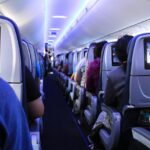 Passengers seated in an aircraft cabin, with rows of seats visible and travellers using personal devices.