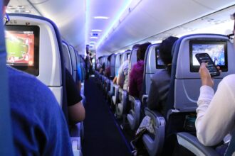 Passengers seated in an aircraft cabin, with rows of seats visible and travellers using personal devices.