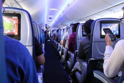 Passengers seated in an aircraft cabin, with rows of seats visible and travellers using personal devices.