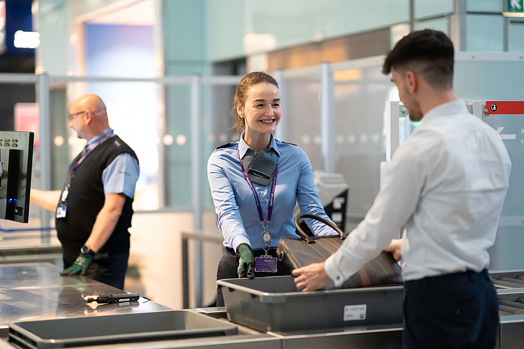 T5 Security officer performing different tasks. Security staff help passengers with checks on personal belongings in trays at a security area in Terminal 5 at London Heathrow Airport.