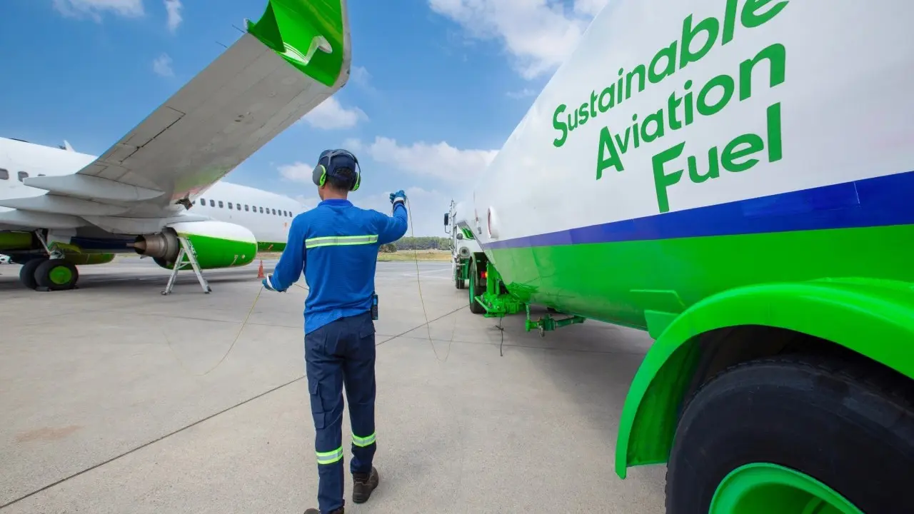 Ground crew member directing a sustainable aviation fuel tanker truck beside a commercial aircraft on the airport apron.