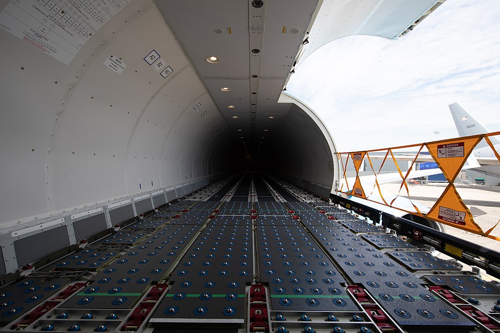 Interior view of an aircraft cargo hold with roller floor panels and the cargo door open.
