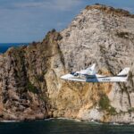 Tecnam aircraft flying in front of rocky cliff, sea below.