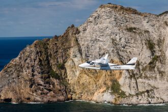 Tecnam aircraft flying in front of rocky cliff, sea below.
