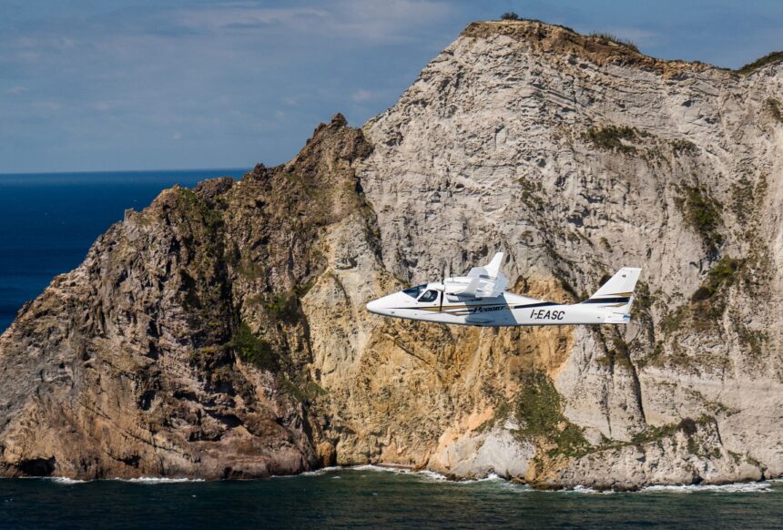 Tecnam aircraft flying in front of rocky cliff, sea below.
