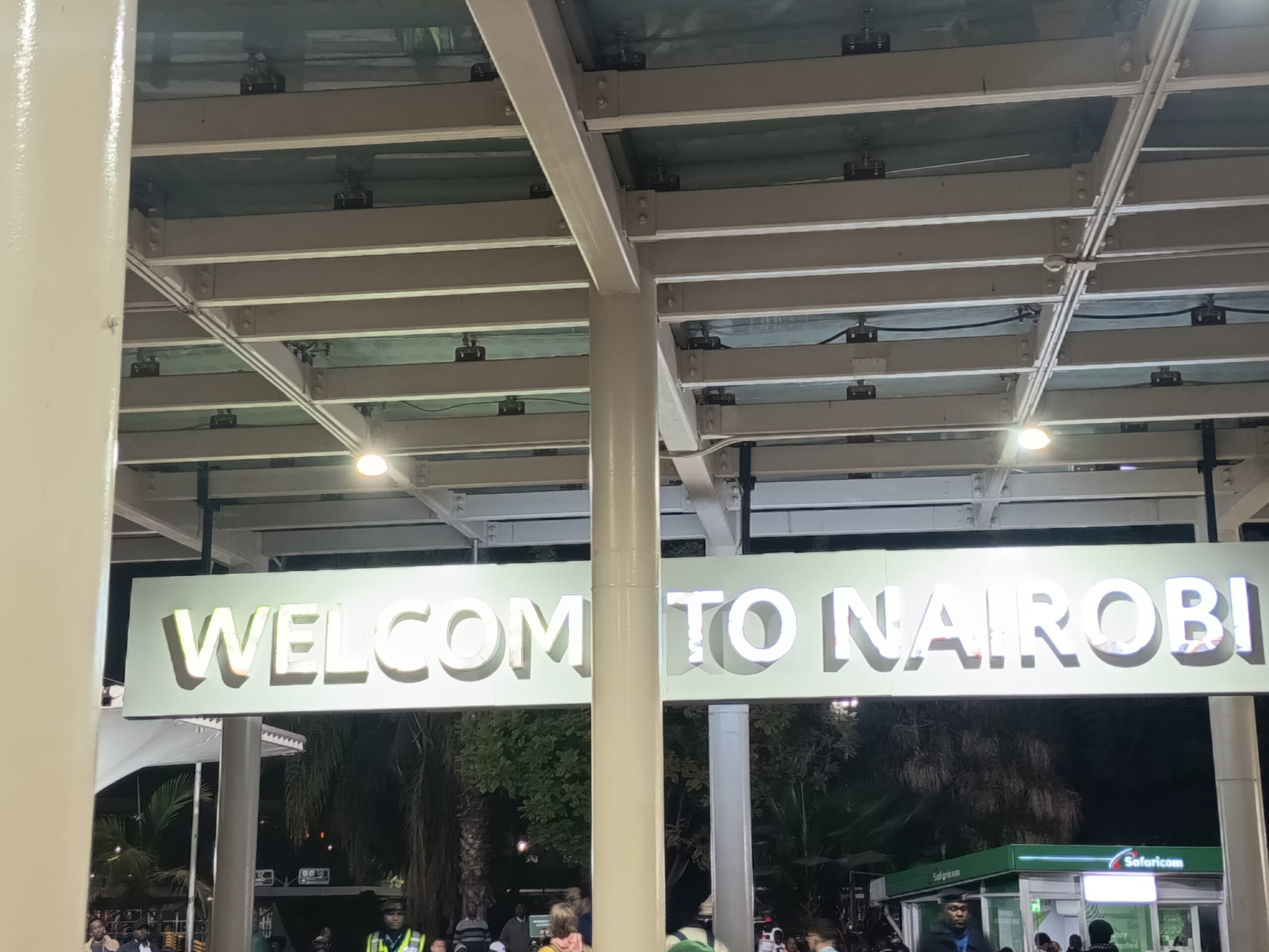 Arrivals at the Jomo Kenyatta International Airport. A sign reads "WELCOME TO NAIROBI". Some people in the image.