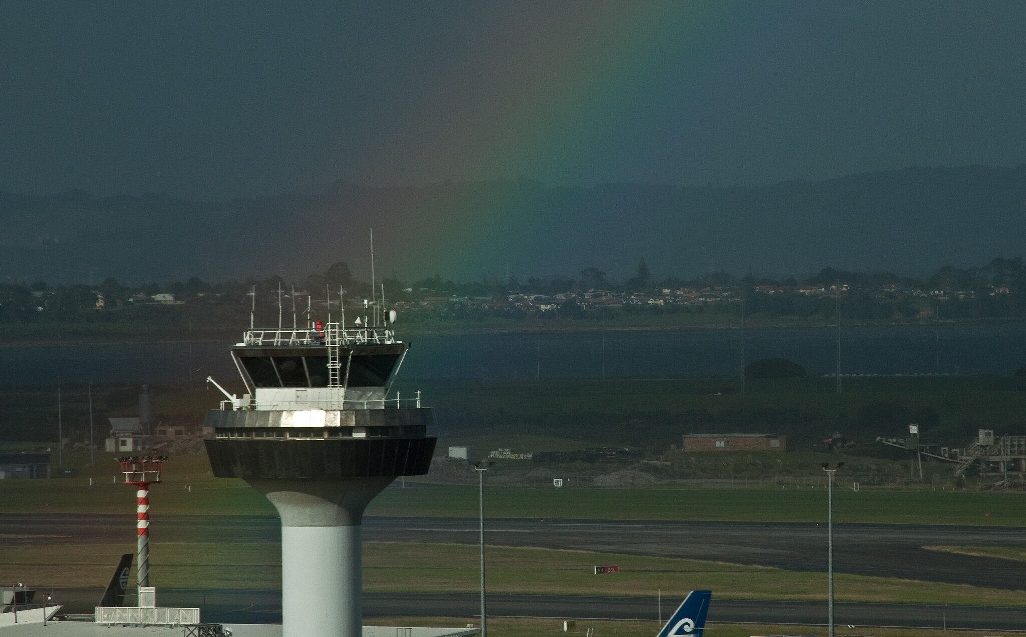 The control tower at Auckland Airport, New Zealand. There is a rainbow behind the tower and an overcast sky.