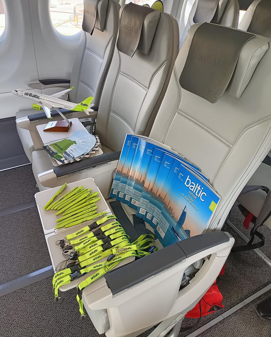 A Business Class cabin showing a stack of brochures, green lanyards, and a scale model of an airBaltic A220-300. 