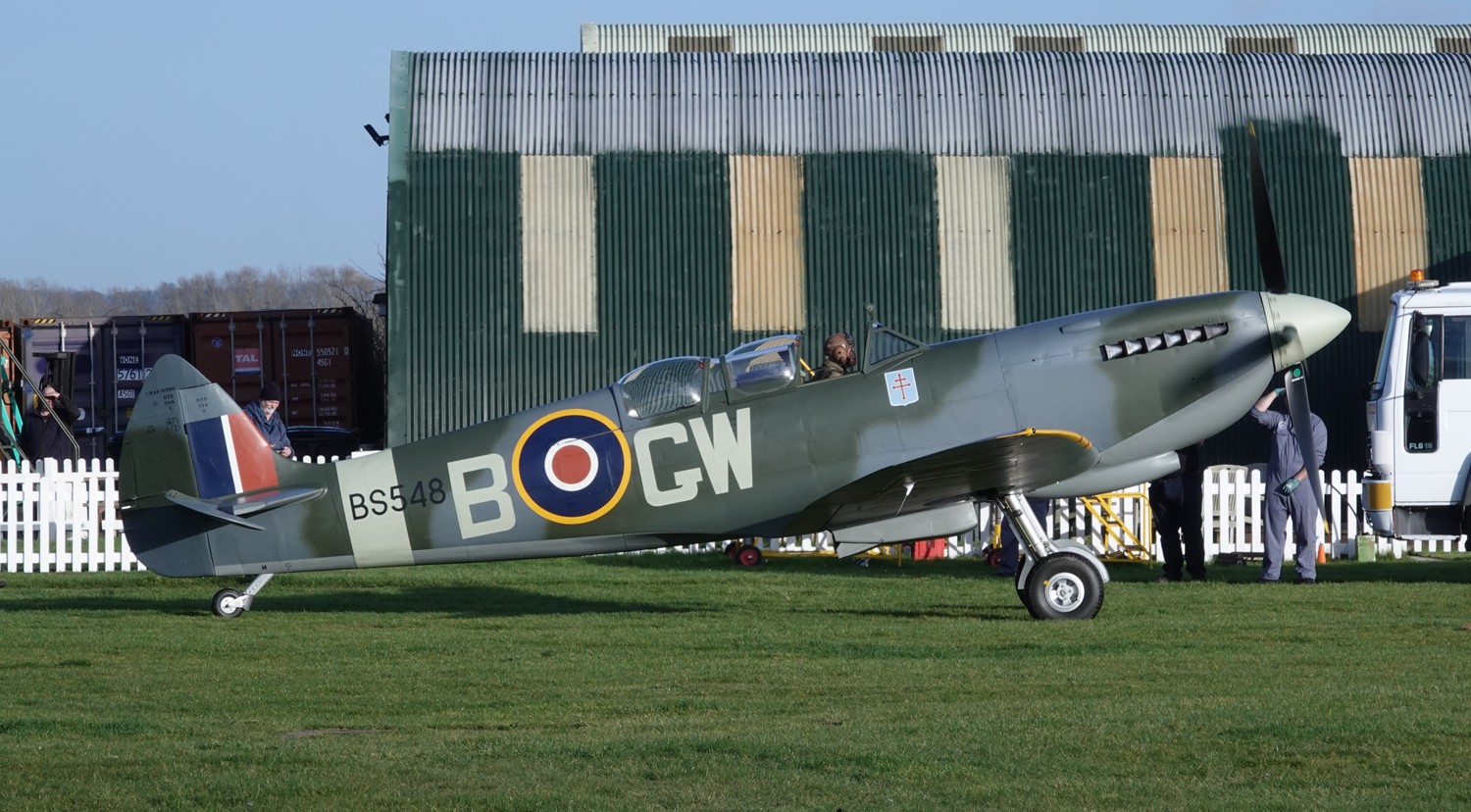 Supermarine Spitfire on the grass with pilot inside, green and grey building behind it.