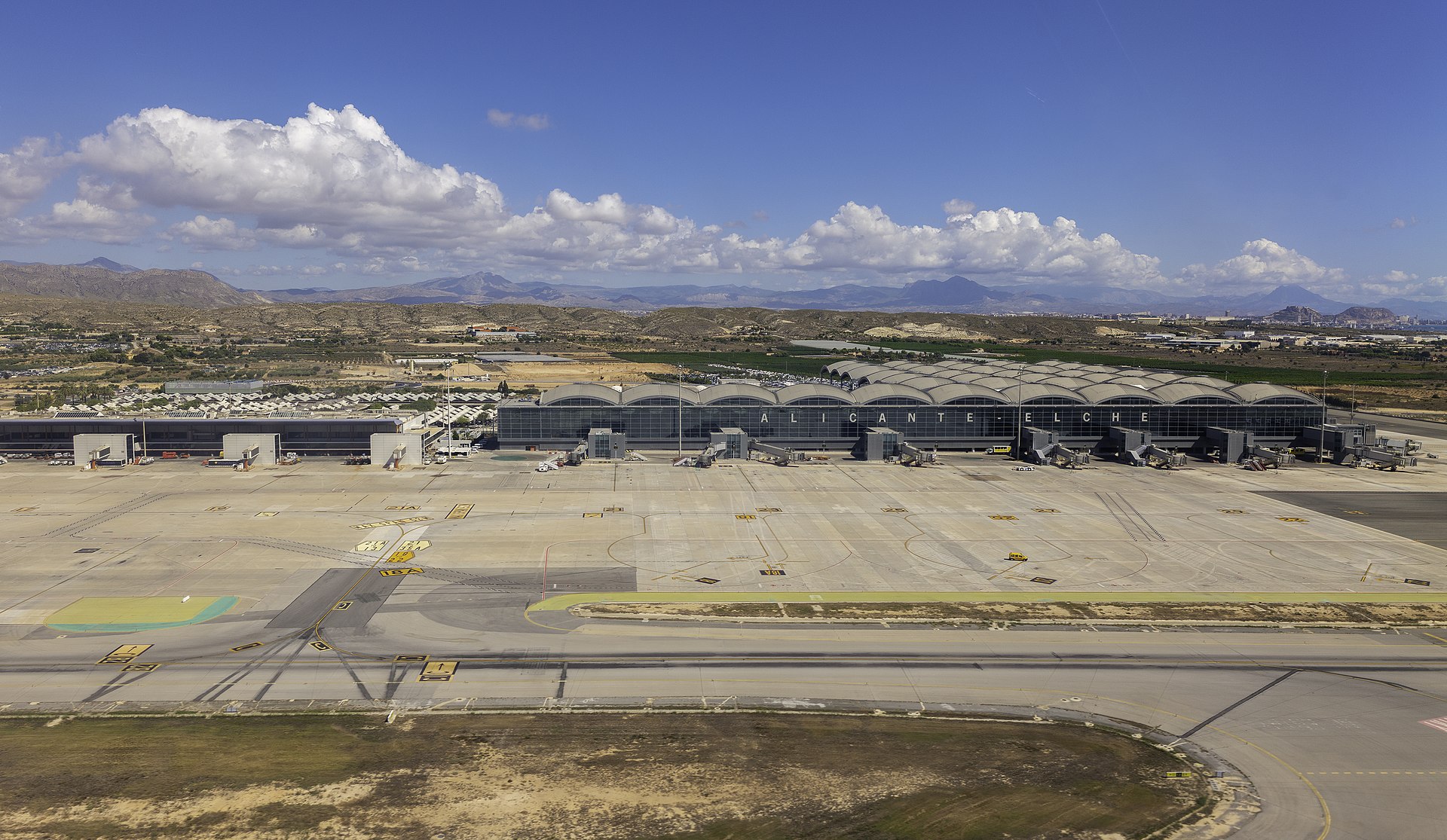 An aerial wide-angle view of the Alicante–Elche Miguel Hernández Airport (ALC) terminal. The terminal features a long, modern glass facade with the name 