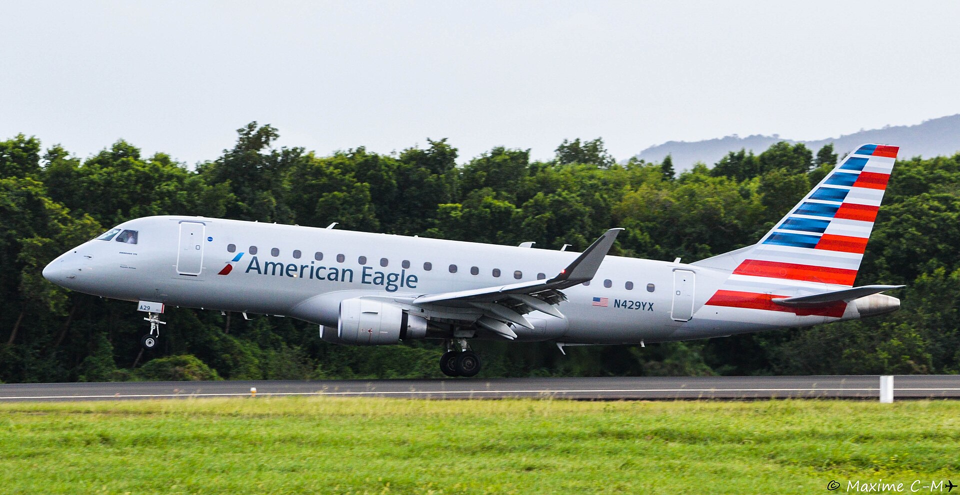Side profile of an American Eagle Embraer 175 regional jet during touchdown or takeoff on an asphalt runway. The aircraft features the iconic red, white, and blue striped tail livery and the tail number N429YX. The background consists of dense, lush green tropical trees under an overcast sky, with a hazy mountain ridge visible in the distance. The foreground shows a grassy airfield.