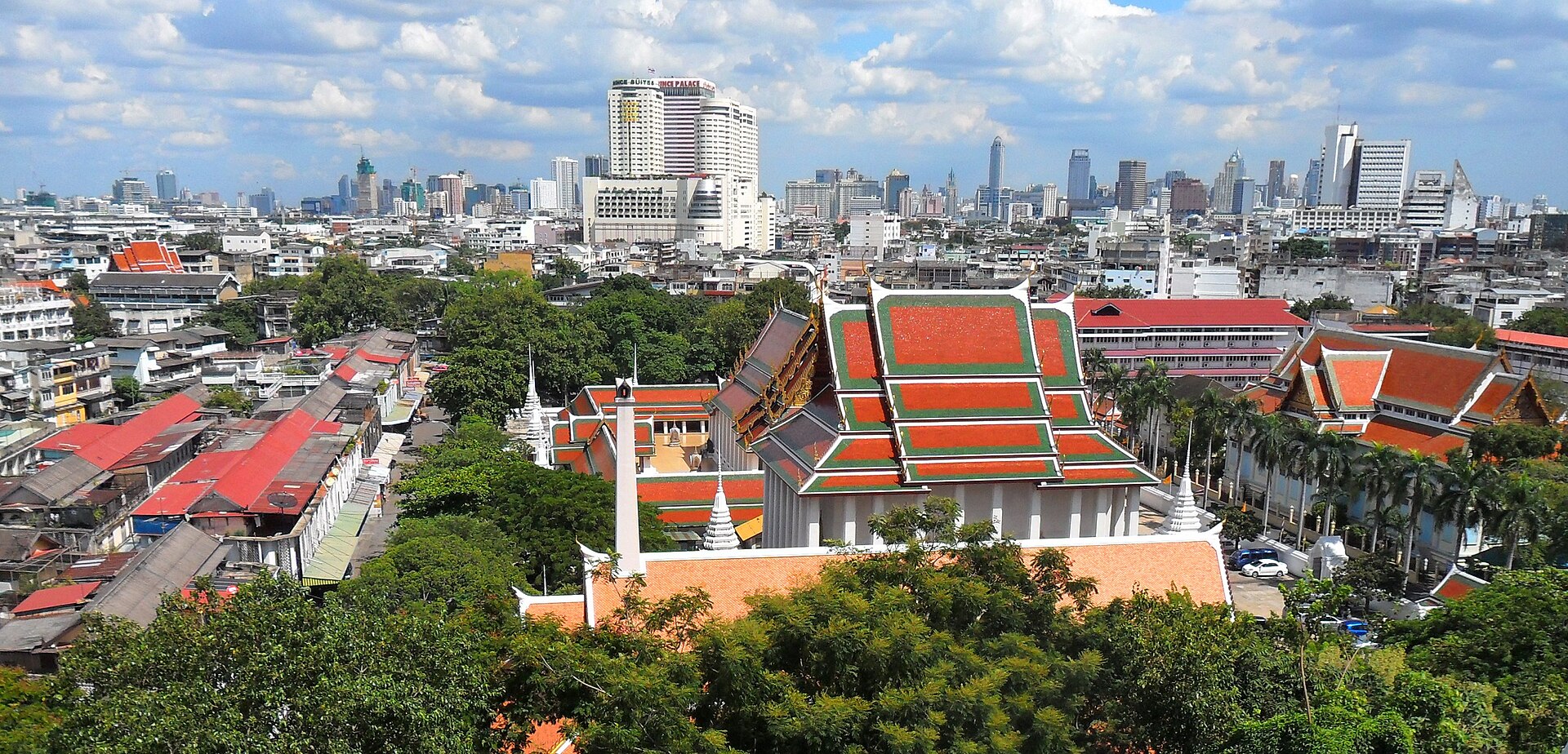 A wide cityscape view of Bangkok, Thailand. In the foreground, traditional buildings with bright orange and green tiled roofs are surrounded by thick green trees. The midground and background feature a dense array of modern white skyscrapers and urban buildings under a blue sky with fluffy white clouds.