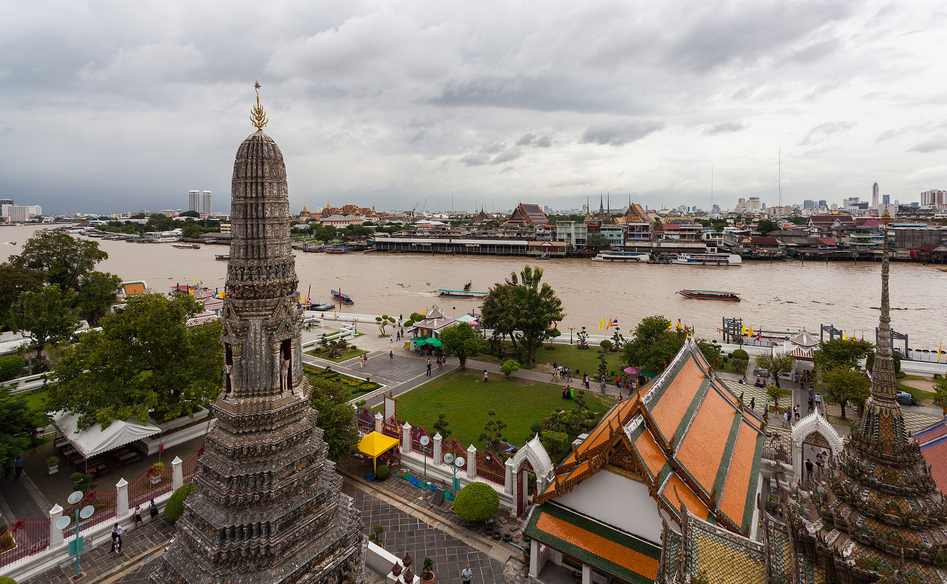 Image shows a high-angle view of the Wat Arun temple complex in Bangkok. A large, ornate grey stone prang (spire) dominates the foreground, with traditional Thai buildings featuring orange-tiled roofs nearby. In the background, the wide, brown Chao Phraya River is filled with longtail boats, with the modern city skyline visible in the distance under a grey, cloudy sky