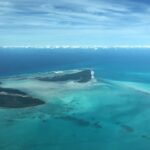 High-altitude aerial photograph of Bimini, Bahamas. Two main tropical islands are separated by a wide expanse of pale, translucent turquoise water and sandbars. The land is covered in dark green vegetation with visible developments and a small airstrip. A line of white fluffy clouds sits low on the horizon under a pale blue, wispy sky, meeting the deep blue edge of the deep ocean.